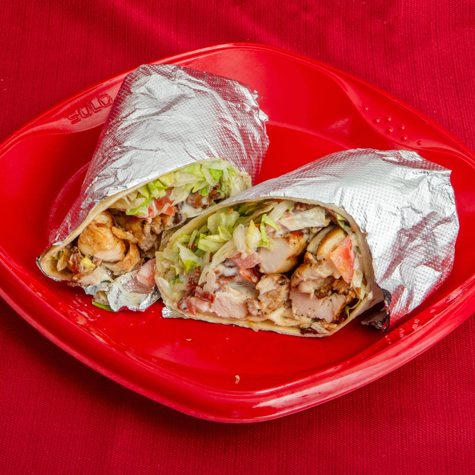 Two foil-wrapped chicken shawarma wraps filled with lettuce, tomatoes, onions, and sauce on a red plate.