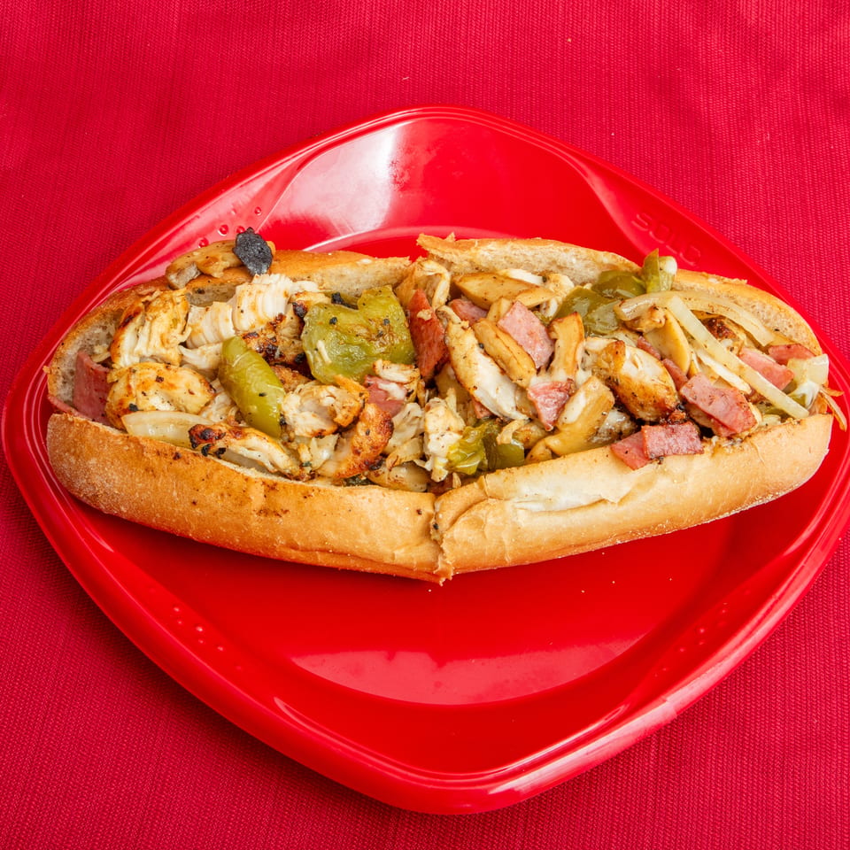 Loaded toasted sub sandwich with grilled chicken, sliced meats, peppers, onions, and mushrooms on a red plate.