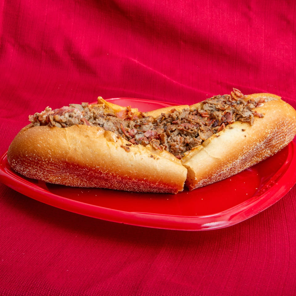 Loaded steak and bacon sandwich on a toasted baguette, served on a red plate against a red fabric background.