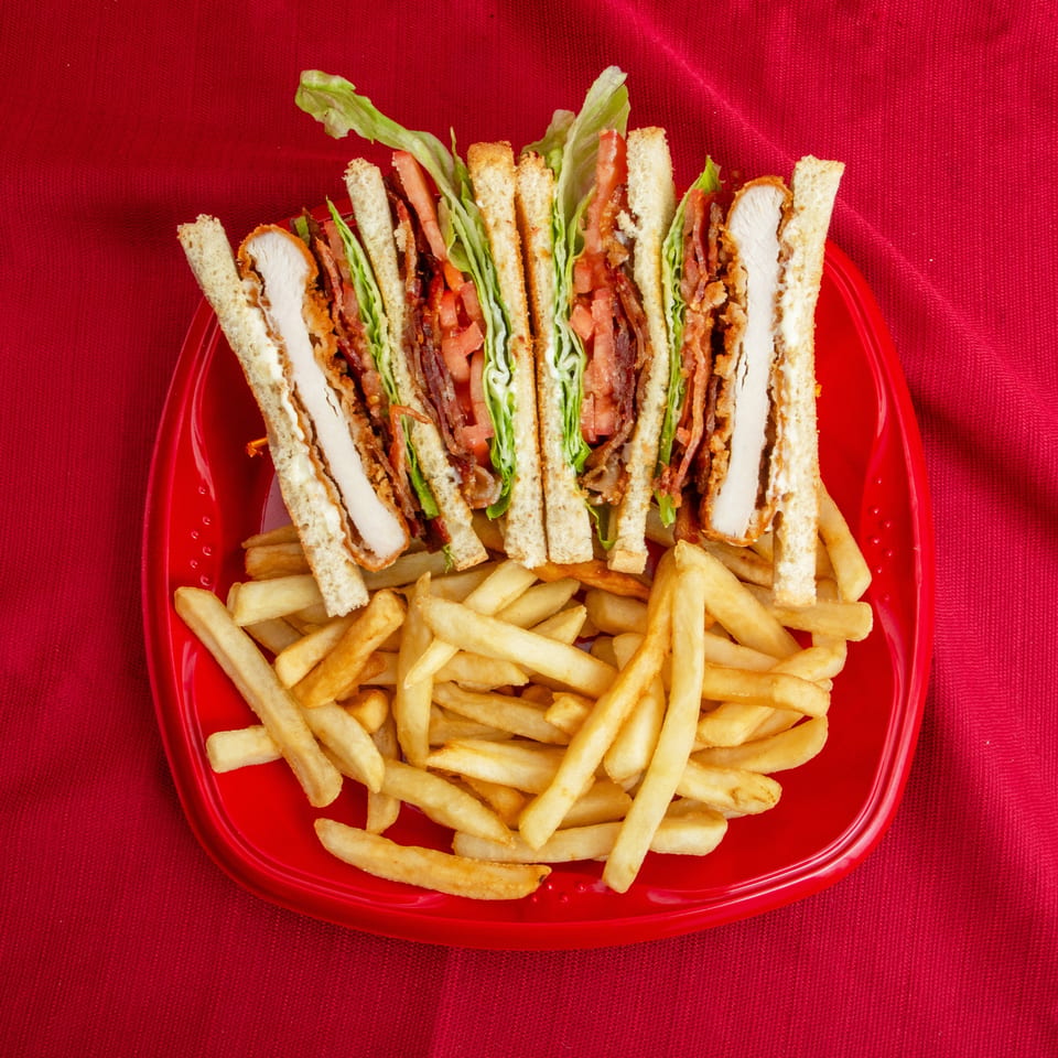 Club sandwich with chicken, bacon, lettuce, and tomato served with French fries on a red plate.